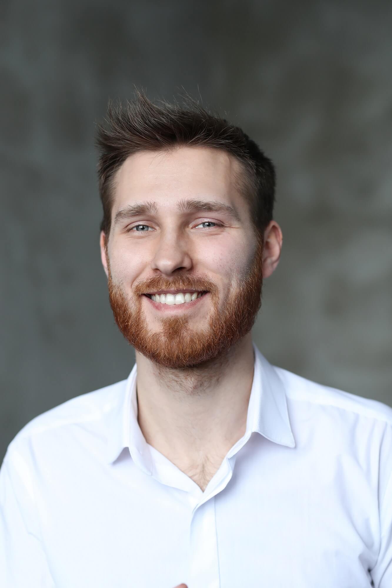A man with a beard and mustache smiles while wearing a white shirt, standing against a blurred gray background.