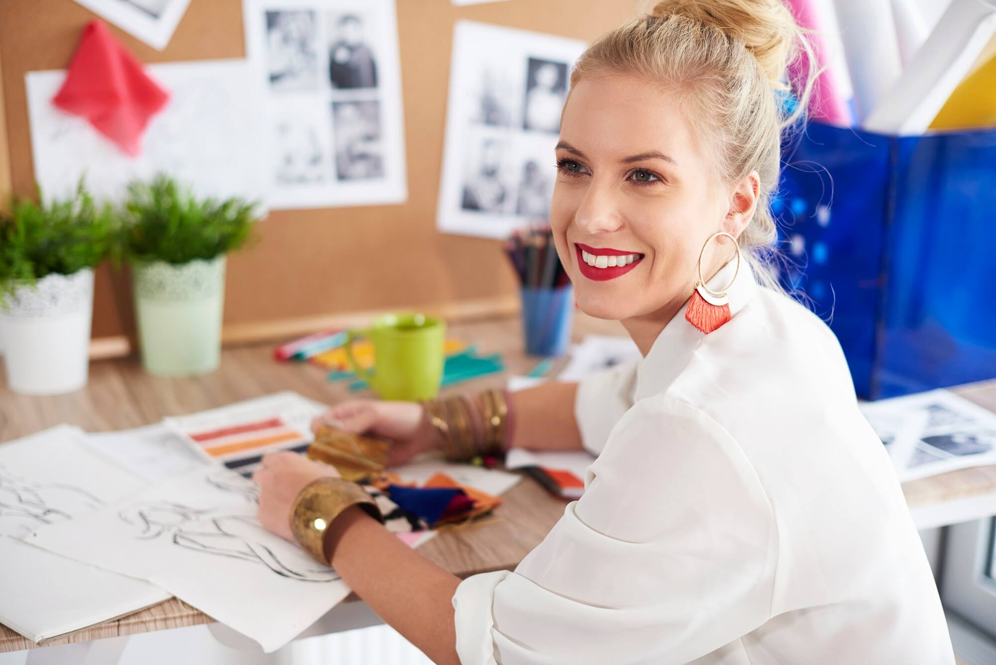 A person with blonde hair and red lipstick sits at a desk with art supplies and sketches, smiling.