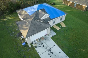 Aerial view of a house, post-storm recovery underway, with a blue tarp covering part of the roof. Shingles and debris are scattered across the driveway and lawn, signs of nature’s recent fury.