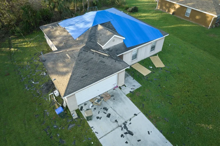 Aerial view of a house, post-storm recovery underway, with a blue tarp covering part of the roof. Shingles and debris are scattered across the driveway and lawn, signs of nature’s recent fury.